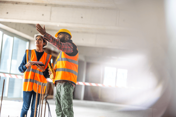 Two construction workers standing in a building undergoing repairs, collaborating on tasks while wearing protective gear