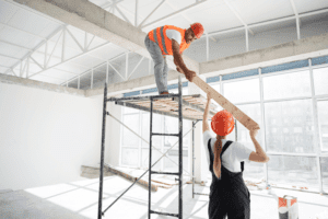 Two construction workers in safety gear handling a timber beam inside a commercial building under renovation.