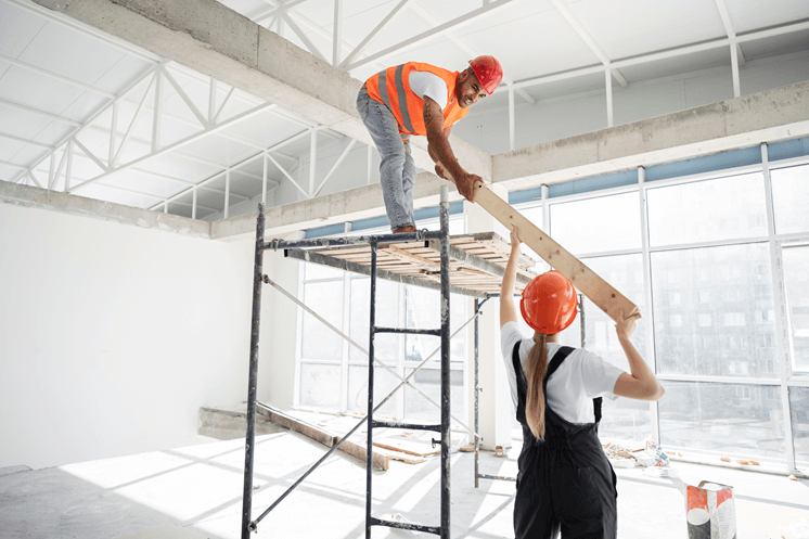 Two construction workers in safety gear handling a timber beam inside a commercial building under renovation.